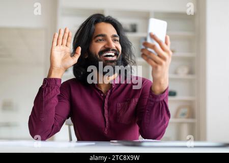Lächelnder indischer Freelancer, der die Hand winkt, während er Videoanrufe auf dem Smartphone hat Stockfoto