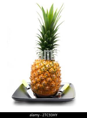 Reife, lebendige Ananas auf einem schwarzen Teller mit Messer und Gabel isoliert über weiß Stockfoto