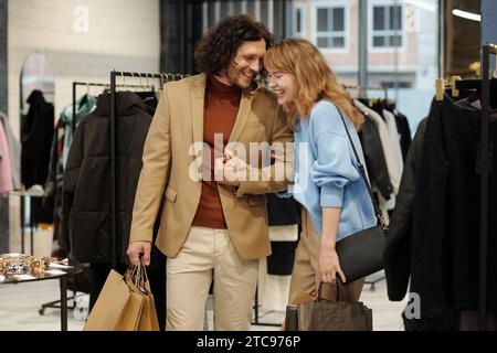 Fröhlicher junger Mann und Frau lachen, während sie in einer Boutique zwischen Regalen mit neuer saisonaler Modekollektion und kleiner Ausstellung stehen Stockfoto