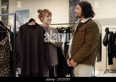 Glücklicher junger männlicher Käufer, der weibliche Angestellte sieht, wie er einen neuen Pullover auf Kleiderbügeln zeigt, während er ihm hilft, Casualwear zu wählen Stockfoto