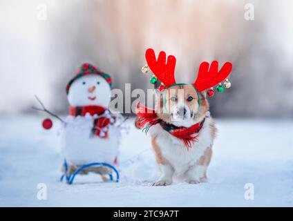 Neujahrskarte mit niedlichem Weihnachtscorgi in Rentiergeweih in einem Schal mit einem Schneemann in einem verschneiten Winterpark Stockfoto