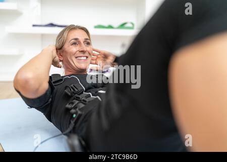 Frau lächelt, während sie Sit-UPS in einem EMS-Trainingsanzug in einem Fitnessstudio macht Stockfoto