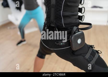 Zwei Frauen, die Squats in einem Fitnessstudio machen, eine trägt einen EMS-Trainingsanzug mit Akku Stockfoto