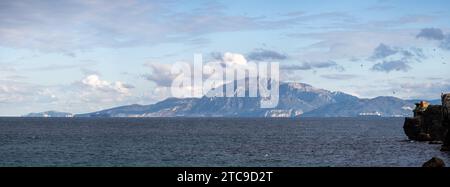 Panoramablick auf ein ruhiges Meer mit einem Berg im Hintergrund unter einem bewölkten Himmel. Stockfoto