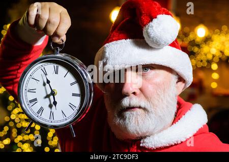 Ernster Weihnachtsmann mit weihnachtlicher Uhr. Zeit zum Feiern. Neujahr oder Weihnachten. Winterfeiertage. Bärtiger Mann mit Weihnachtsmann-Hut mit Stockfoto