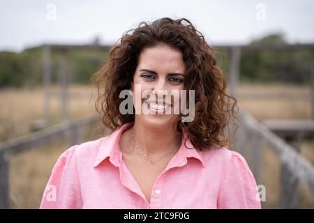 Eine junge weißbrünette Frau, welliges Haar und eine rosa Bluse, die die Kamera lächelnd mit einem Hintergrund eines Naturparks betrachtet. Kaukasische Frau Stockfoto