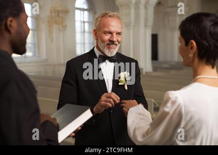 Porträt eines lächelnden älteren Bräutigams, der während der Hochzeitszeremonie am Kirchenaltar Ringe mit seiner Frau austauscht Stockfoto