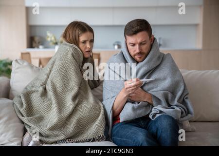 Familienpaar junger Mann, Frau, die Temperatur misst, sitzt auf der Couch und ist in Plaids eingewickelt. Stockfoto