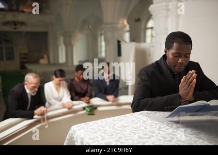 Porträt eines jungen afroamerikanischen Priesters, der am Altar mit einer Gruppe von Menschen während des Sonntagsgottesdienstes in der Kirche betet Stockfoto