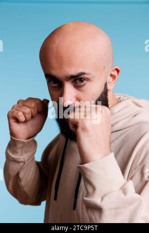 Arabischer Mann zeigt Stärke, während er mit geballten Fäusten in Boxposition steht Porträt. Junge Person, die eine Kampfgeste macht, während sie mit aggressivem Gesichtsausdruck in die Kamera schaut Stockfoto