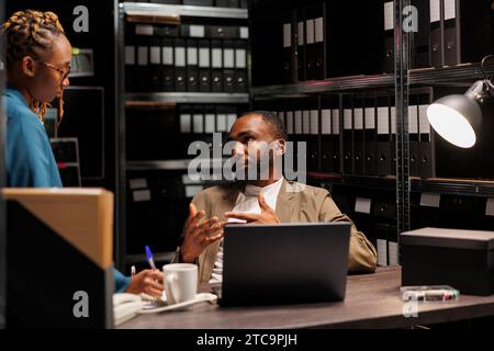 Ermittler von Männern und Frauen, die in einem dunklen Raum voller Regale mit Mappen ein Brainstorming und eine Diskussion über Kriminalermittlungen durchführen. afroamerikanische Detektive studieren Berichte mit Archivakten und Laptop Stockfoto