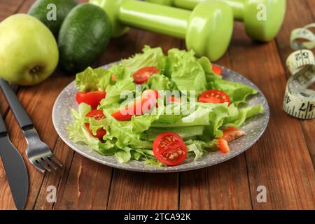 Gesunde Ernährung. Salat, Besteck, Kurzhanteln und Maßband auf Holztisch, Nahaufnahme Stockfoto