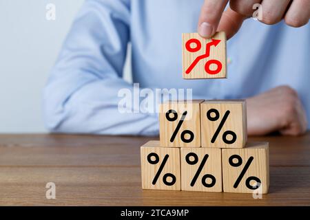 Steigende Hypothekenzinsen, dargestellt durch Prozentzeichen mit Aufwärtspfeil. Mann baut eine Pyramide aus Würfeln am Holztisch, Nahaufnahme Stockfoto