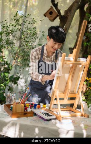 Junger glücklicher asiatischer Künstler in einer Schürze färbt sich auf einer Leinwand auf einer Staffelei in seinem Garten. Kunstwerke, Kunstworkshop, kreative Aktivitäten Stockfoto