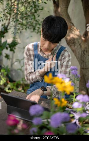 Ein junger asiatischer Mann, Besitzer eines kleinen Blumenladens, spricht am Telefon mit seinem Kunden, während er an einem Tisch in seinem Blumengarten arbeitet. Stockfoto