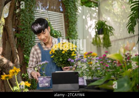 Ein glücklicher und gutaussehender junger asiatischer Blumenhändler oder Gärtner arbeitet in einer Pflanzenzüchtung in einem Garten. Florist, Gärtner, Blumenhändler, Unternehmer Stockfoto