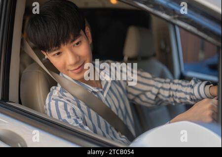 Ein glücklicher und gutaussehender junger asiatischer Mann im lässigen Outfit fährt ein Auto und schaut auf den Seitenspiegel. Stockfoto