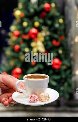 Eine Hand hält eine Tasse türkischen Kaffee und türkische Delikatesse oder Lokoum vor einem geschmückten Weihnachtsbaum in Istanbul, Türkei Stockfoto