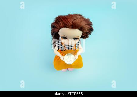 Eine kleine Kinderpuppe mit einer Münze in den Händen auf blauem Hintergrund. Stockfoto