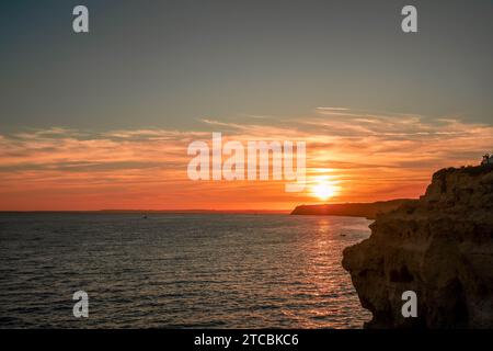 Ein ruhiger Sonnenuntergang auf einer felsigen Klippe mit Blick auf den Ozean, der das warme Leuchten der untergehenden Sonne gegen den tieforange Himmel einfängt Stockfoto