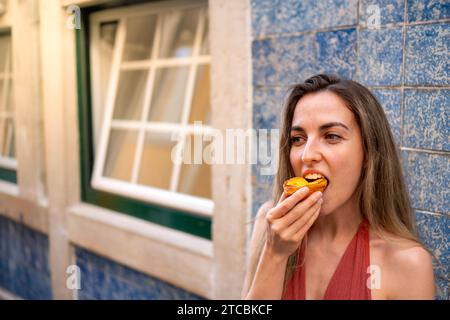 Nahaufnahme, Frontalansicht einer jungen Frau, die ein traditionelles portugiesisches Pastel de Nata genießt, horizontal bei Tageslicht aufgenommen Stockfoto