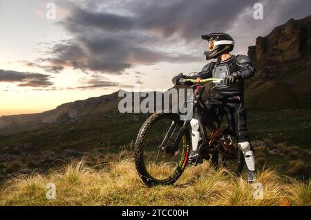 Der Fahrer in vollem Schutz auf einem Mountainbike steht auf einem Hügel und betrachtet den Sonnenuntergang auf dem Hintergrund der Felsen im Kaukasus Stockfoto