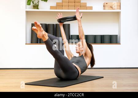 Seitenansicht einer schlanken Frau in Sportbekleidung, die wegschaut, während sie auf einer Matte liegt, mit einem magischen Kreis in erhobenen Händen im Fitnessraum und die Beine gegen die Wand streckt Stockfoto