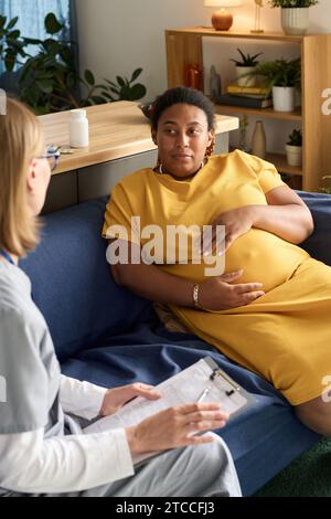 Vertikales Bild einer afroamerikanischen schwangeren Frau, die mit der Krankenschwester spricht, während sie Medizin verschreibt Stockfoto