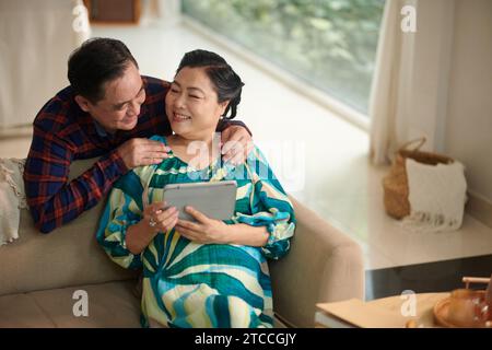 Fröhlicher älterer Mann, der seine Frau umarmt und auf einem Tablet-Computer eine Show ansieht Stockfoto