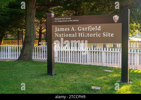 Ein braunes Parkschild mit der Aufschrift James A. Garfield National Historic Site steht auf dem Rasen vor einem weißen Lattenzaun und hohen Bäumen in Ohio. Stockfoto