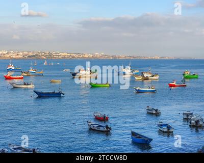 Küste des Atlantischen Ozeans, kleine Bucht mit vielen bunten Fischerbooten neben dem Yachthafen von Cascais und in der Ferne auf der Klippe sichtbare Stadtlandschaft. Stockfoto