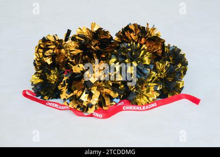 Goldene und schwarze Pompons mit einem roten Cheerleading-Band auf einer weißen Übungsmatte Stockfoto