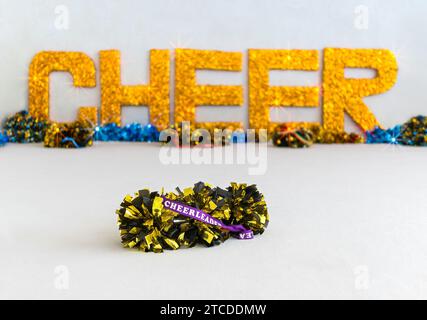 Goldene und schwarze Pompons mit „Cheerleader“ auf einem lila Band und großen goldenen, glitzernden „CHEER“-Buchstaben im Hintergrund Stockfoto