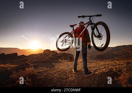 Männlicher Mountainbiker geht bergauf und trägt ein Fahrrad auf dem Rücken zum Sonnenuntergang Berg Stockfoto