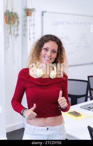 Frau mit Kopfhörern, die Daumen nach oben zeigen und lächeln, während sie in ihrem Büro posiert. Geschäfts- und Erfolgskonzept. Stockfoto