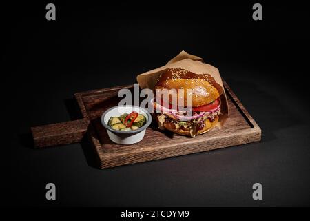 Köstlicher Hamburger mit Sesambrötchen, Rindfleisch und Gurken als Beilage auf schwarzem Holztablett Stockfoto