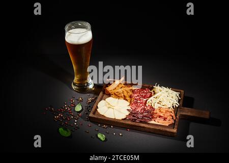 Ein Glas kaltes Bier in der Nähe der Wurstplatte mit Gourmet-Käse, getrocknetem Rindfleisch und Salami auf schwarzem Hintergrund Stockfoto