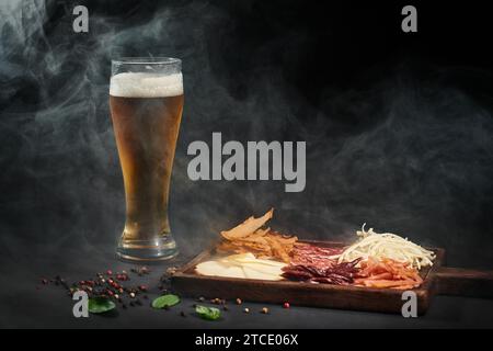 Ein Glas Bier in der Nähe einer Wurstplatte mit Gourmet-Käse, getrocknetem Rindfleisch und Salami vor schwarzem Hintergrund Stockfoto