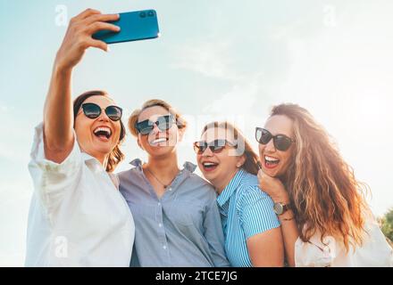 Porträt von vier fröhlichen, lächelnden Frauen in einer Sonnenbrille, die sich zusammenschließen und Selfie-Fotos mit einem modernen Smartphone machen, während sie im Freien spazieren gehen. Die der Frau Stockfoto