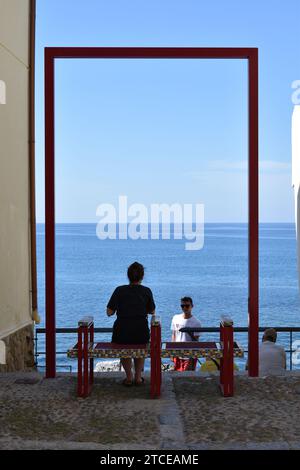 Frauen sitzen auf einer Bank unter einer großen, rechteckigen Kunstinstallation mit Blick auf das Mittelmeer Stockfoto