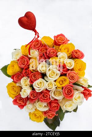Buntes Blumenstrauß von Rosenblüten aus mehrfarbigen Rosen in drei Farben, auf weißem Schneehintergrund. Rotes Souvenir-Herz ragt aus einem Blumenstrauß hervor. Valen Stockfoto