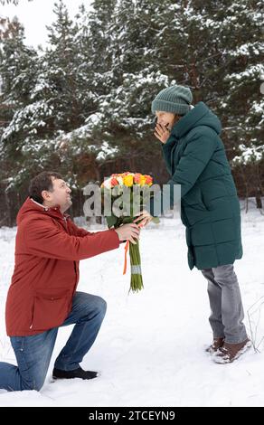 Verliebtes Paar im Winter im Wald oder Park. Ein Mann steht auf einem Knie und schenkt einer bewundernden Frau einen großen Rosenstrauß. Valentinstag. Romantische R Stockfoto