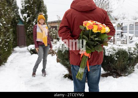Der Mann versteckt einen großen Rosenstrauß hinter seinem Rücken, und eine glückliche Frau steht vor ihm im Hintergrund. Valentinstag. Romantische Relationshi Stockfoto