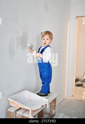 Kleiner Bauarbeiter, der die Wand in einer Wohnung streicht, die gerade renoviert wird. Ein niedliches Kind im Arbeitsanzug, das Malerwalze benutzt, während es zukünftig zu Hause spielt. Stockfoto