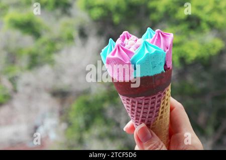 Hand hält einen rosa und blauen Eiskegel mit verschwommenem grünem Laub im Hintergrund Stockfoto