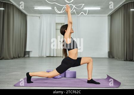 Schlanke afroamerikanische Tänzerin im Sportbekleidungstraining in Kriegerstellung auf der Fitnessmatte im Tanzsaal Stockfoto