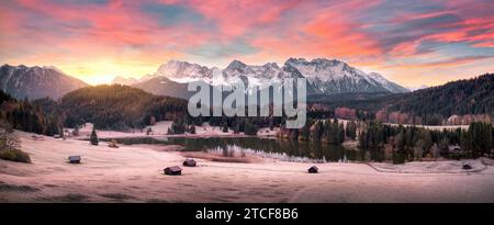 Atemberaubende Landschaft bei Sonnenaufgang mit Bergen, einem malerischen See und Wäldern auf Hügeln in den Alpen, gefrorene Wiese und Hütten, wunderschöne Panoramaaufnahme der Natur Stockfoto