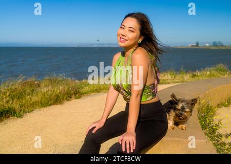 Wind geweht, schöne kurvige athletische asiatische Frau und Yorkie Dog, die in der Nähe der San Francisco Bay sitzen Stockfoto