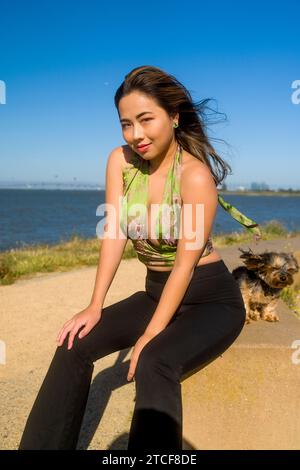 Wind geweht, schöne kurvige athletische asiatische Frau und Yorkie Dog, die in der Nähe der San Francisco Bay sitzen Stockfoto