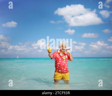 Mann, der im Meer steht, eine Zigarre raucht und ein Glas Cocktail hält Stockfoto